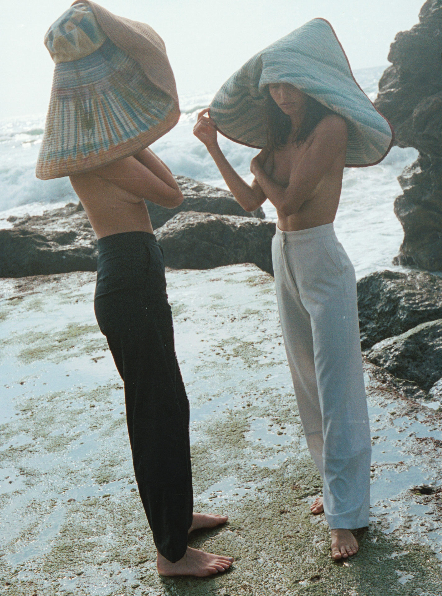2 femmes en pantalons, les pieds dans l'eau sur la plage portant des chapeaux en forme de coquillages