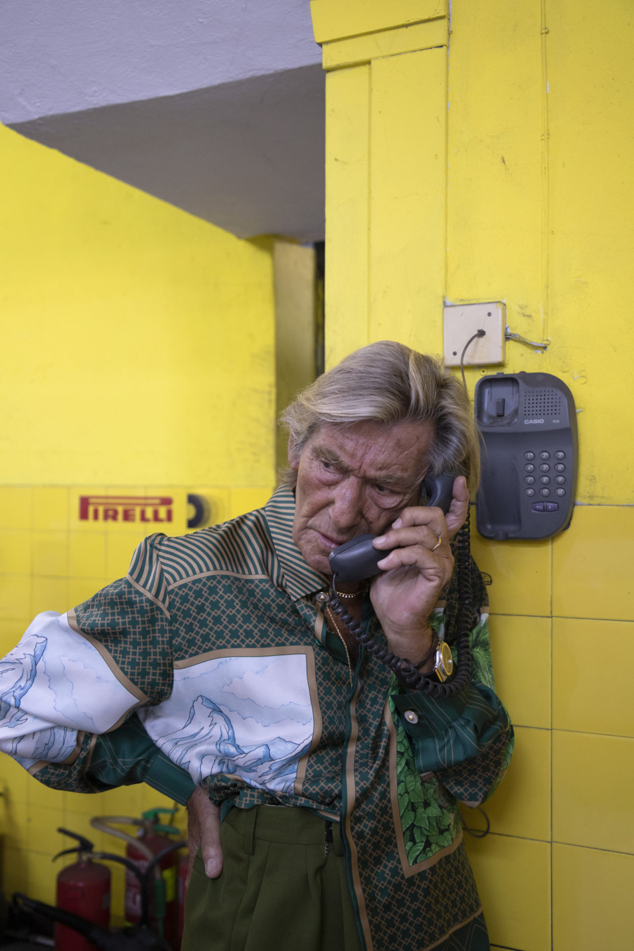 Homme agé en veste verte et blanche téléphonant d'un téléphone publique dans une salle de carrelage jaune