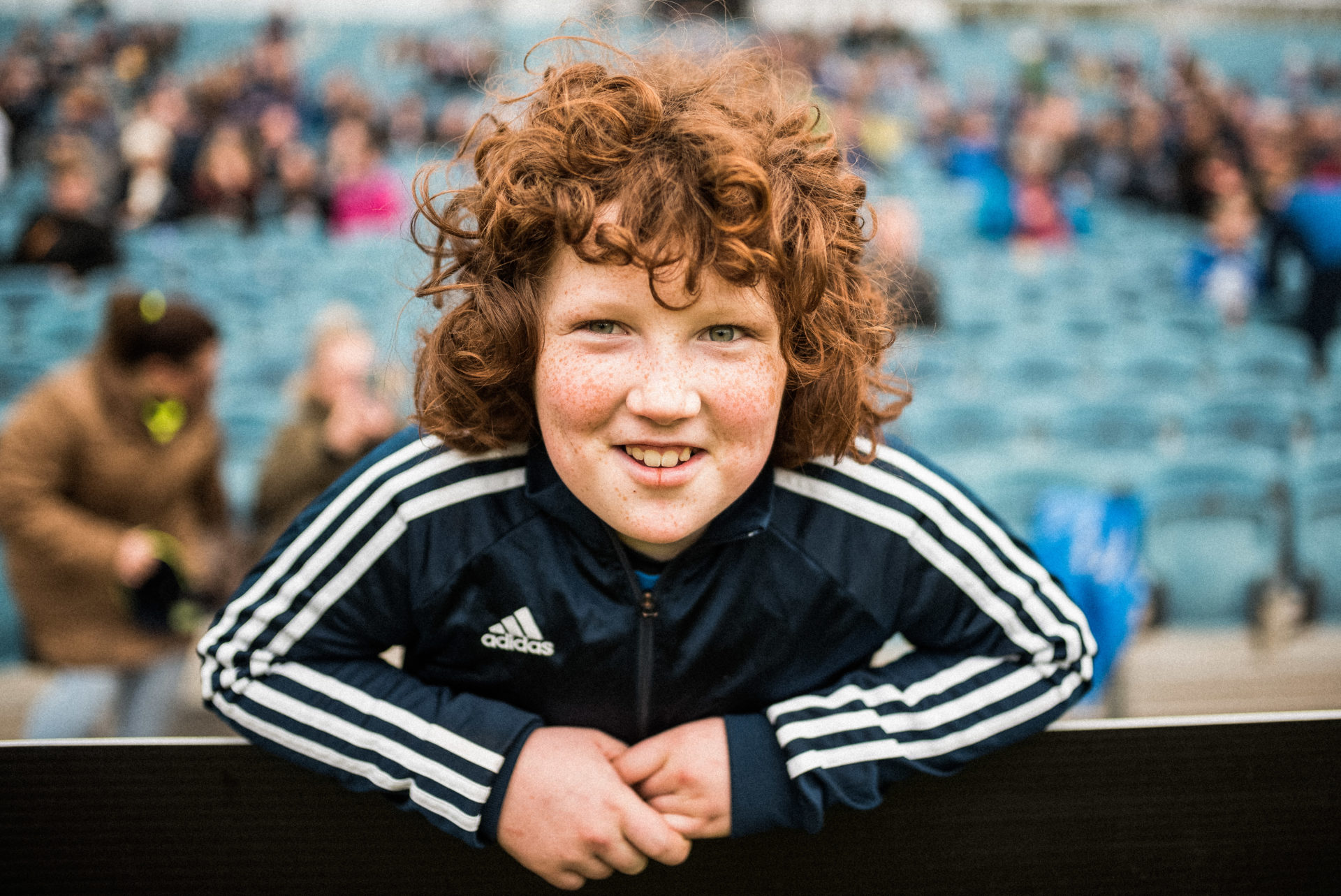 Portrait d'un enfant au cheveux roux frisé et en tenue de sport adidas, souriant dans les gradins d'un stade