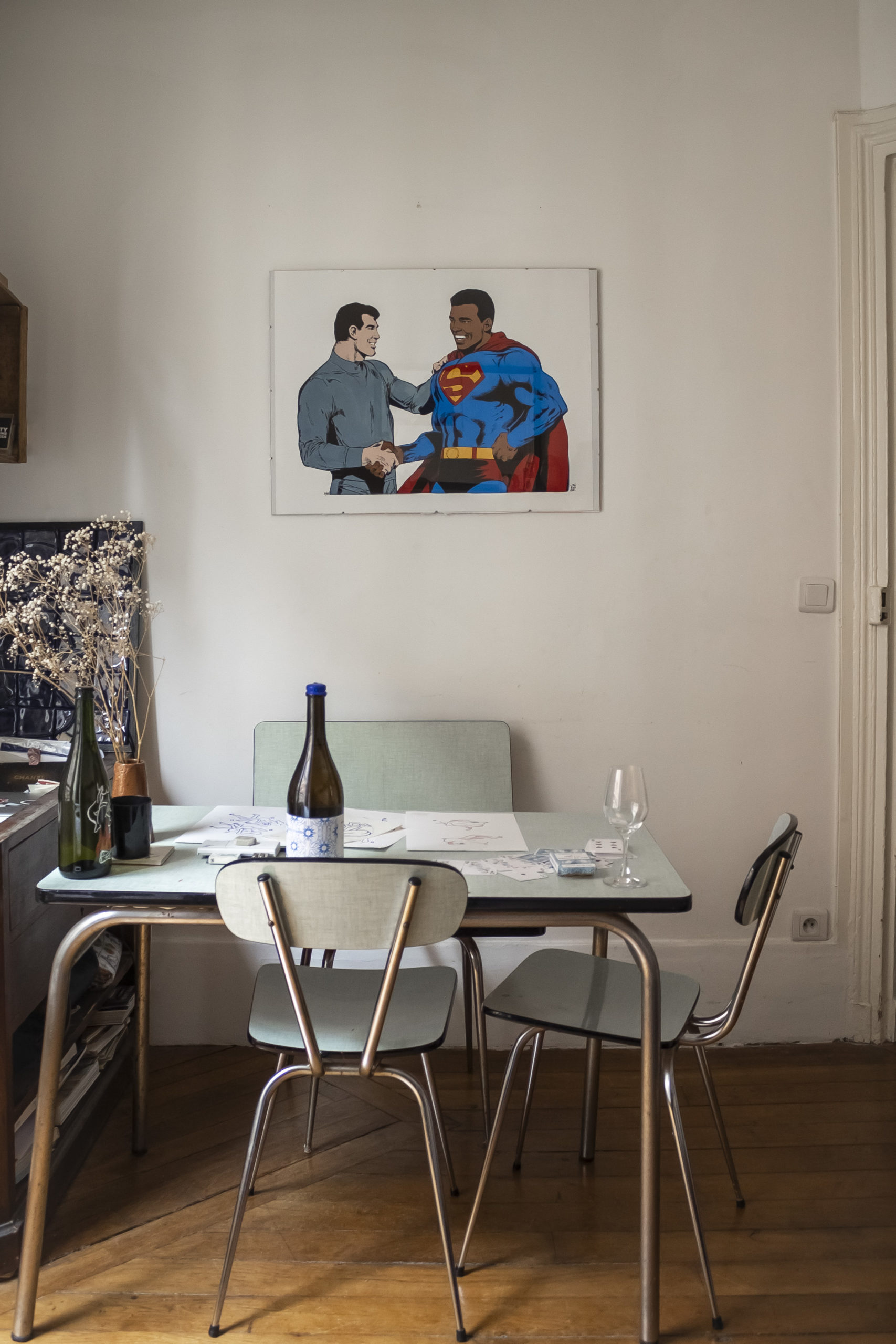 Vue de la table en formica bleu de Karla Sutra positionnée sour un cadre représentant un superman noir. Sur la table sont posés des dessins, une bouteille de vin et des verres