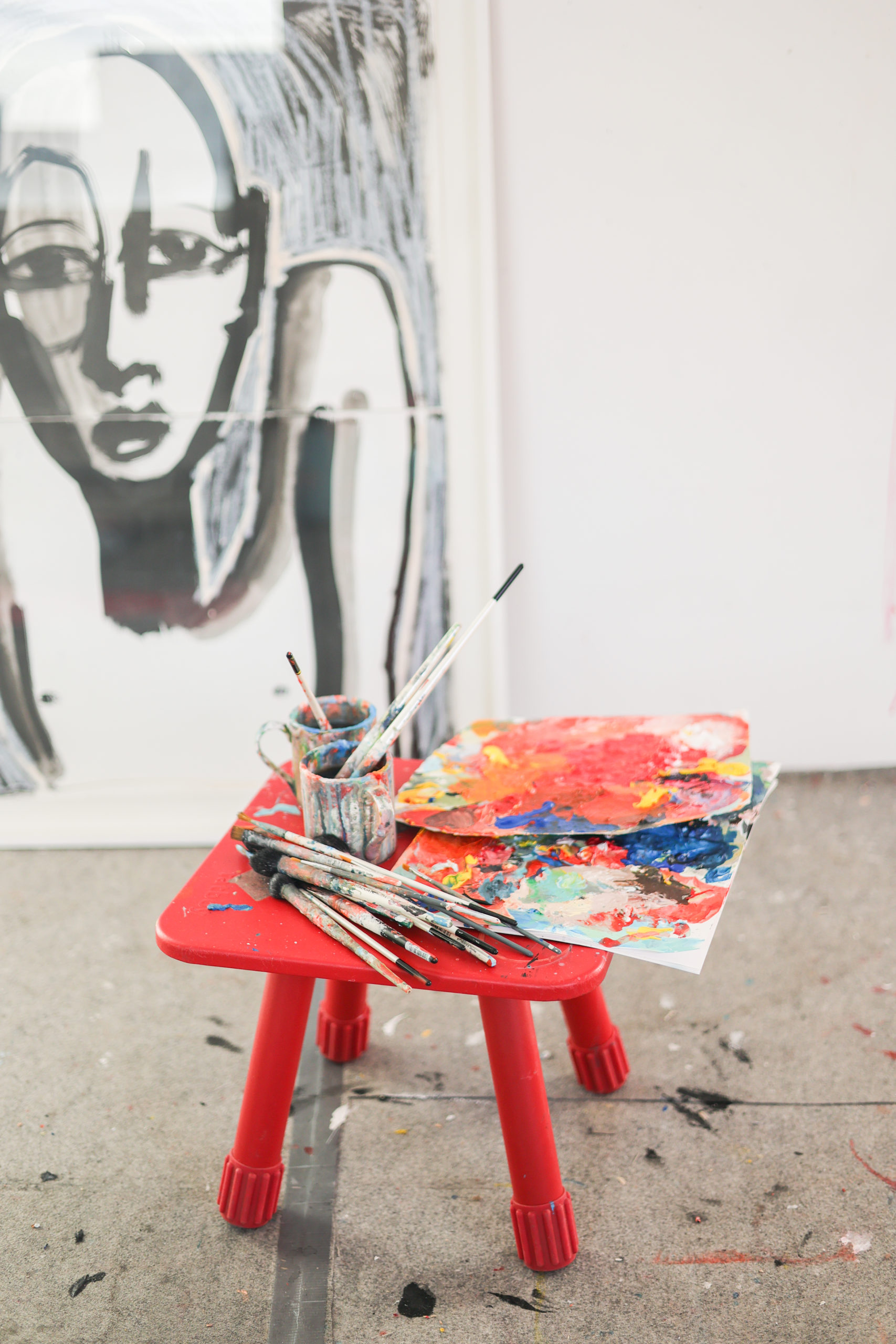 Une palette de peinture et des pinceaux sont posés sur un petit tabouret rouge. Une peinture dans les tons gris est posé en arrière plan