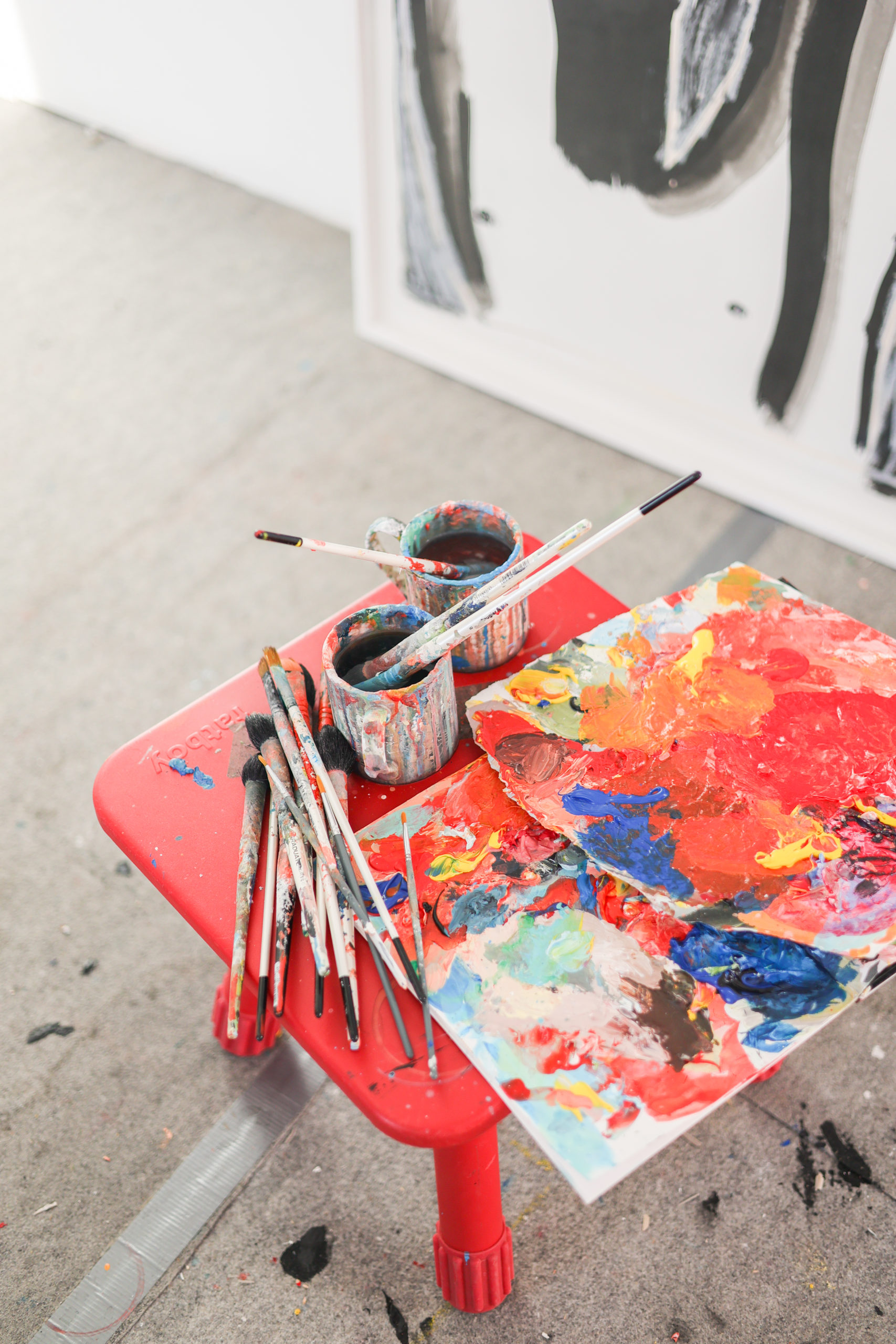Zoom sur une palette de peinture, des pinceaux et des pots posés sur un tabouret en plastique rouge
