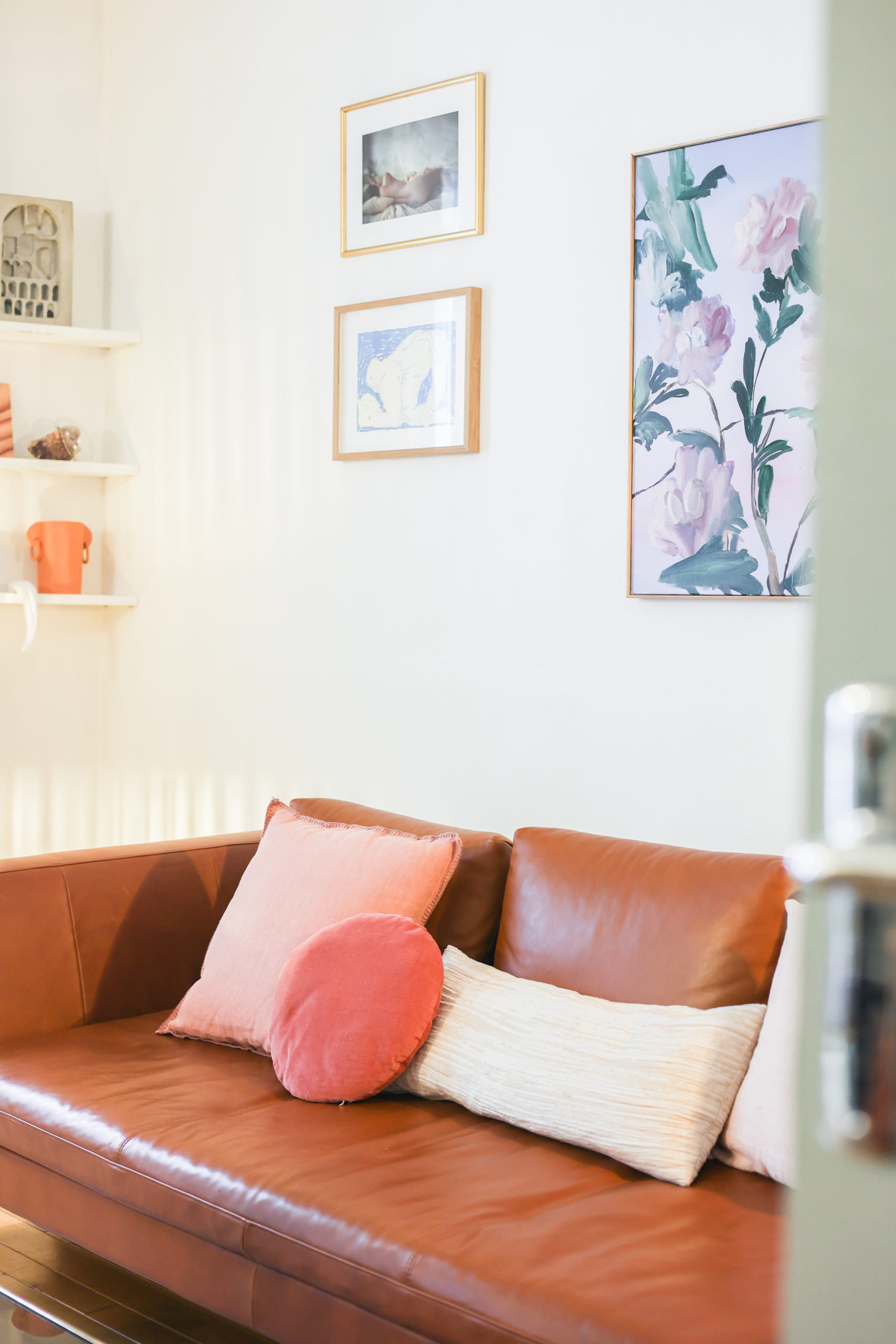 Canapé en cuir marron, avec des coussins roses posé devant un mur avec 3 cadres 