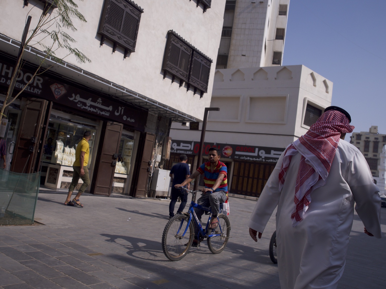 Un homme à vélo à Djeddah devant des immeubles blanc avec en premier plan un homme prtant un qamis