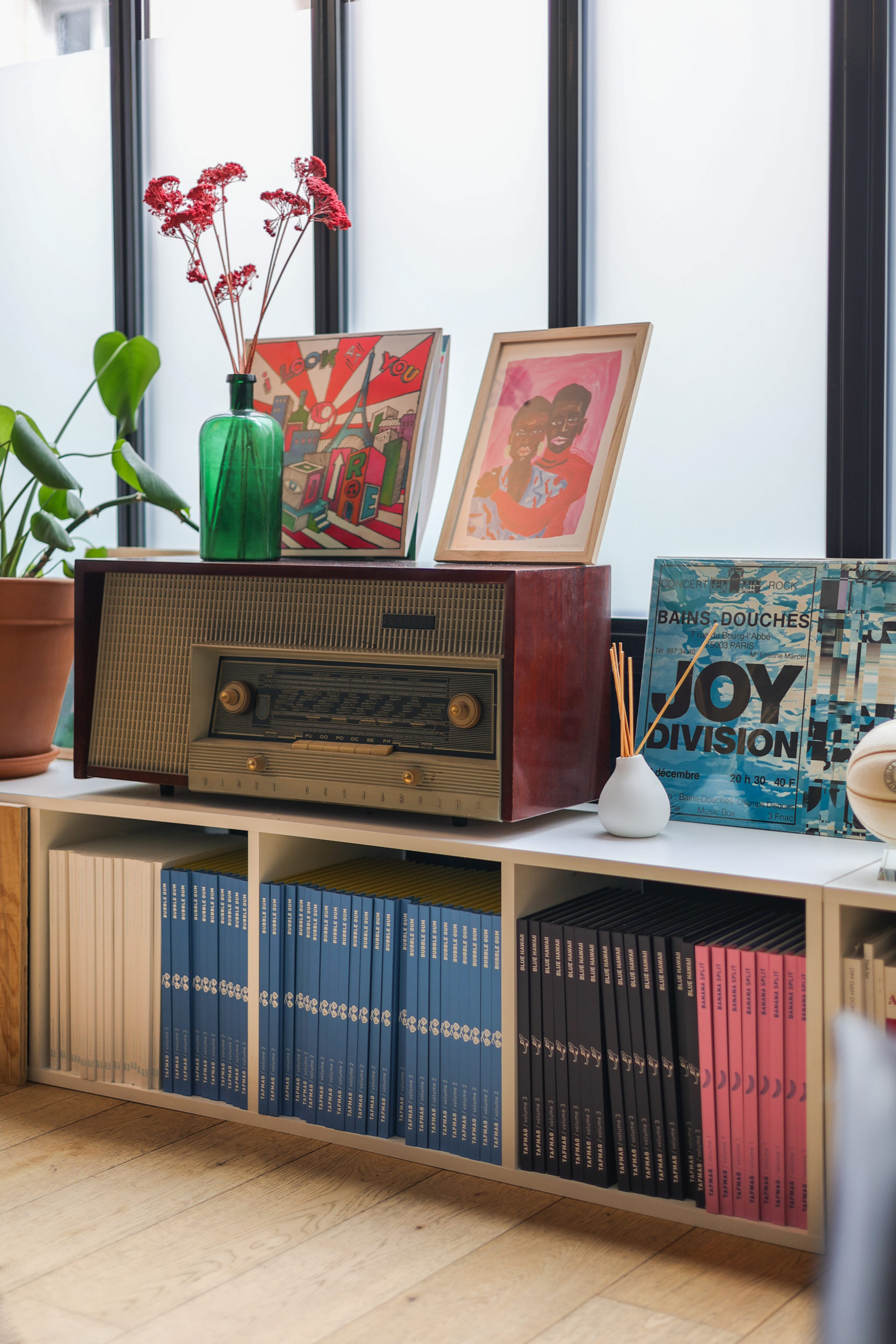 La collection de livre Tafmag dans une étagère blanche sur laquelle sont disposés une radio vintage, des cadres et des vinyles