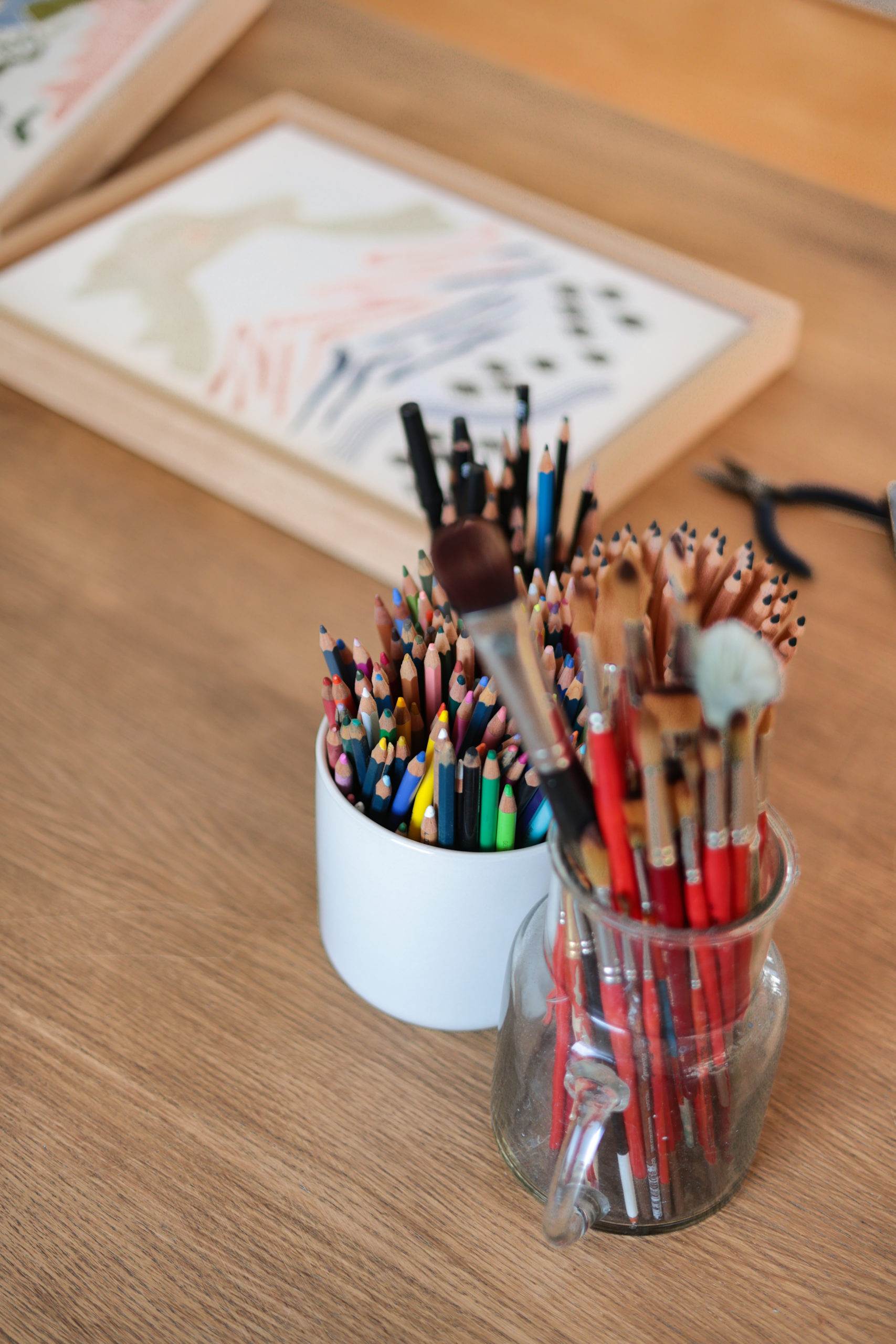 pots contenant les crayons de couleurs et pinceaux de Célia Bruneau posé sur une table en bois à côté d'une broderie